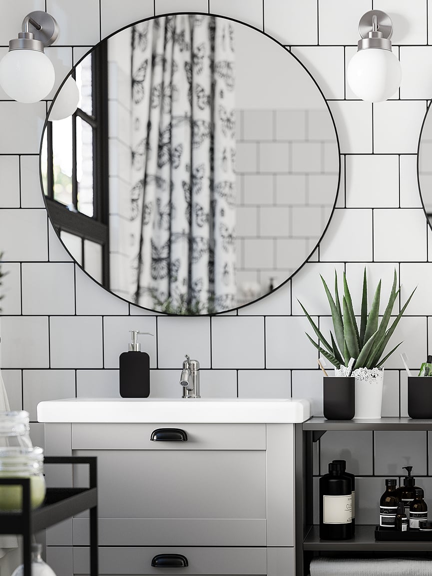 Bathroom with tiled wall, round mirror with two wall lamps, a wash-stand, a potted plant, multiple bottles, and  a trolley.