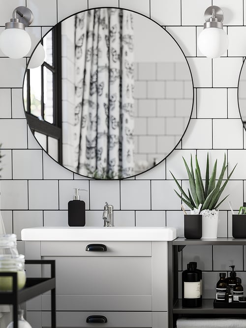 Łazienka ze ścianą wyłożoną płytkami ceramicznymi, okrągłym lustrem, dwiema lampami ściennymi, szafką z umywalką, rośliną w doniczce, kilkoma butelkami oraz wózkiem.
