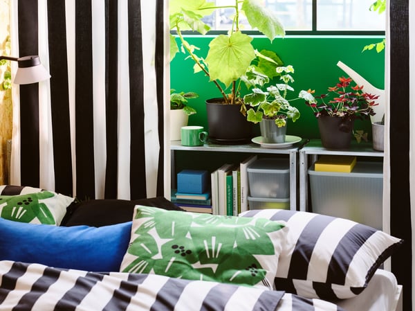 A bed with black/white/striped SLÅNHÖSTMAL bed linen in front of HYLLIS shelving units with green plants on top.