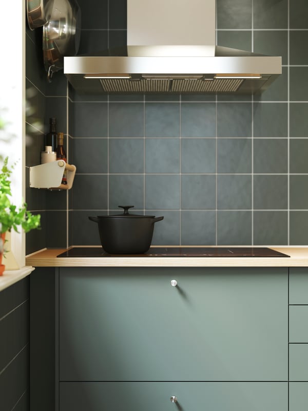 A green-tiled kitchen with NICKEBO fronts in matte grey-green with a hood, a black hob and a cast iron pot on top of it.
