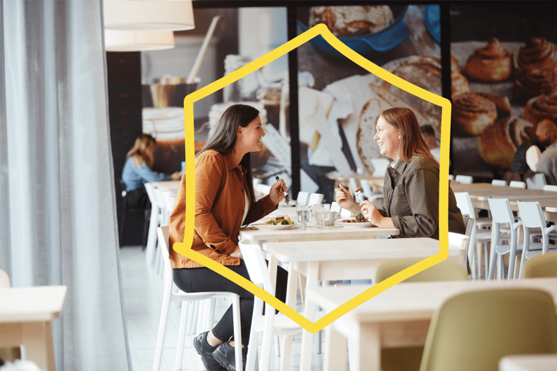 Zwei Frauen sitzen lachend in einem IKEA Restaurant und sind dabei etwas zu essen und zu trinken. 