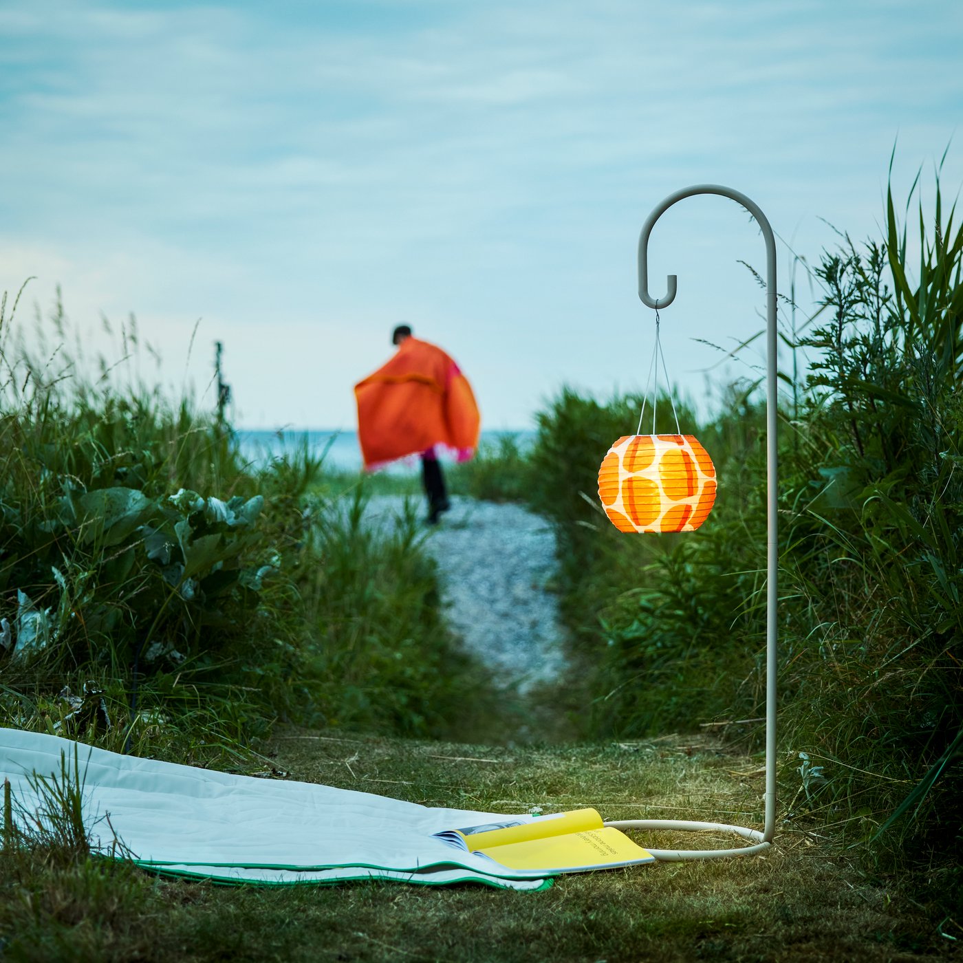 A garden evening setting features a beige SOLVINDEN lamp stand with an illuminated orange lampshade and a person walking.