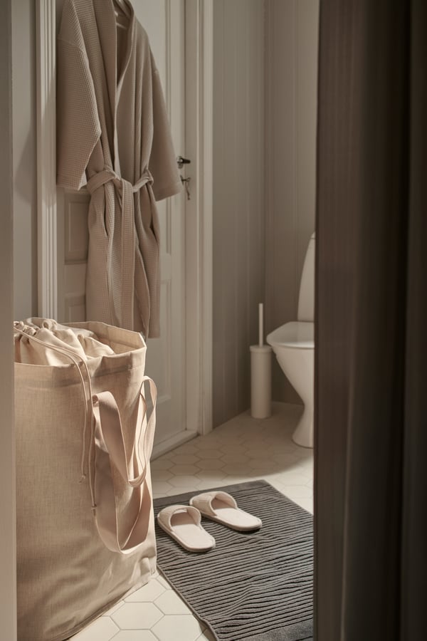A side view of a bathroom with a beige PURRPINGLA laundry bag and a beige robe hanging off a hook.