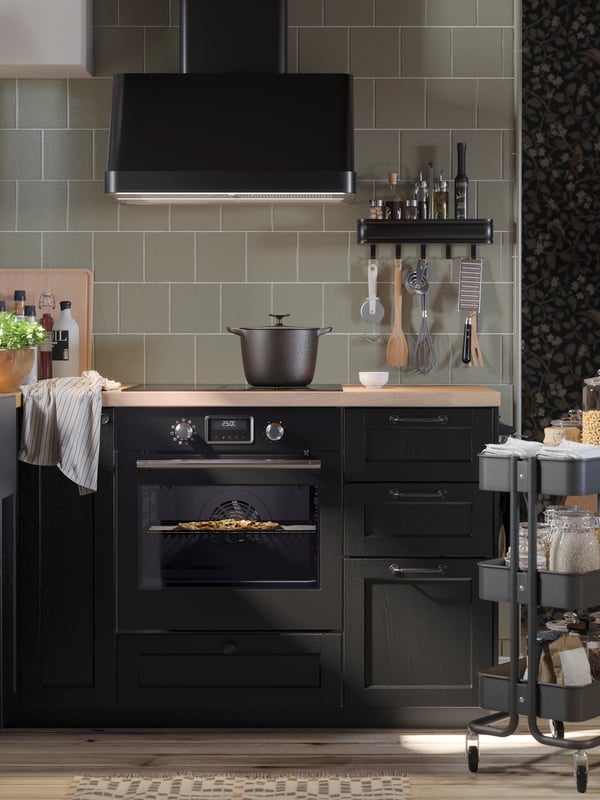 Una cocina con azulejos verdes con frentes LERHYTTAN acabados en negro y electrodomésticos negros, con una placa de inducción, un horno y una campana extractora.