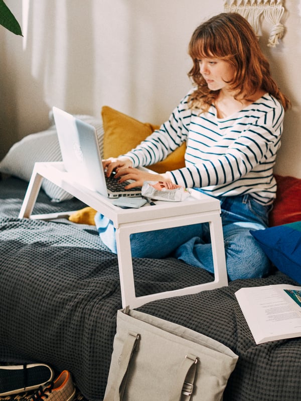 A girl with long red hair and a striped jumper sits on a bed and writes on a laptop placed on a white KLIPSK bed tray.