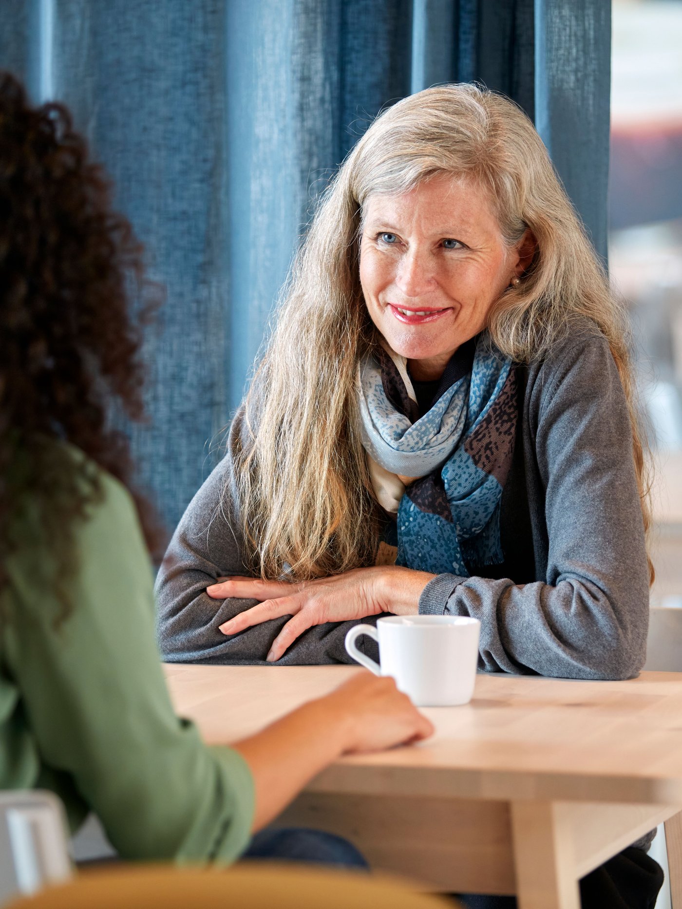 En kvinde sidder og smiler til en anden kvinde foran hende står en hvid kaffekop og de sidder ved et bord.