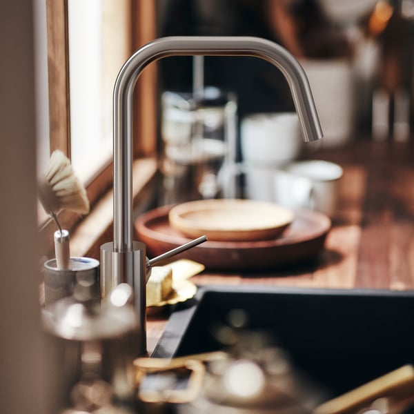 A close-up of a ÄLMAREN kitchen tap above a sink with dishes abd cups in the background.
