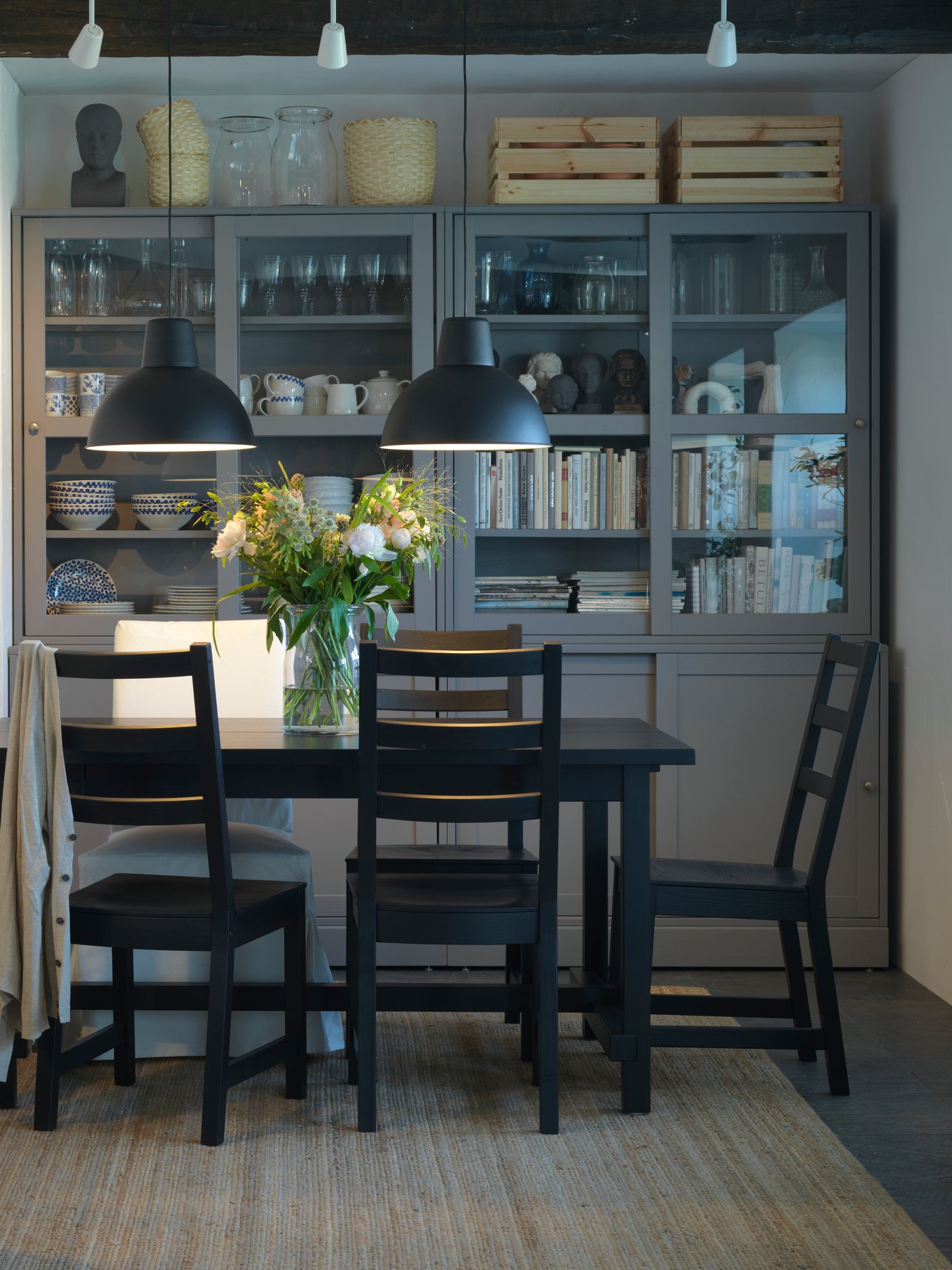 Comedor con mesa y sillas negras, tapete de yute, lámparas de techo negras y combinación de almacenaje con puertas correderas.