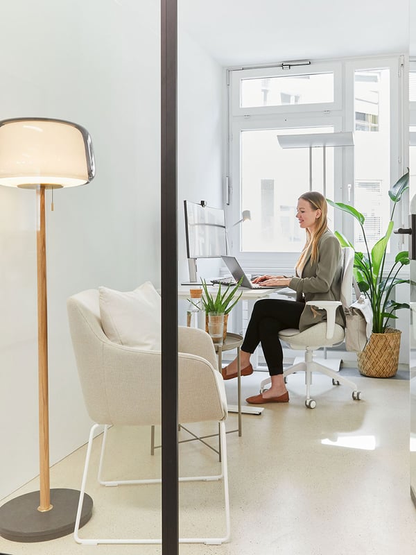 Dans un bureau avec une façade en verre, une femme est assise à un bureau devant la fenêtre. A côté d'elle se trouve une plante en pot et devant elle un fauteuil pour les visiteurs avec une table d'appoint et derrière elle un lampadaire en verre avec un pied en bois et en marbre. 