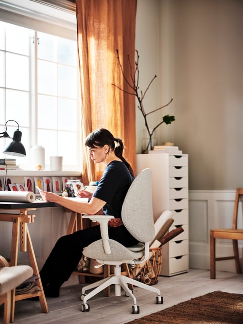 Workspace area featuring a person sitting in an office chair with armrests, a drawer unit, a desk, a work lamp, and a basket.