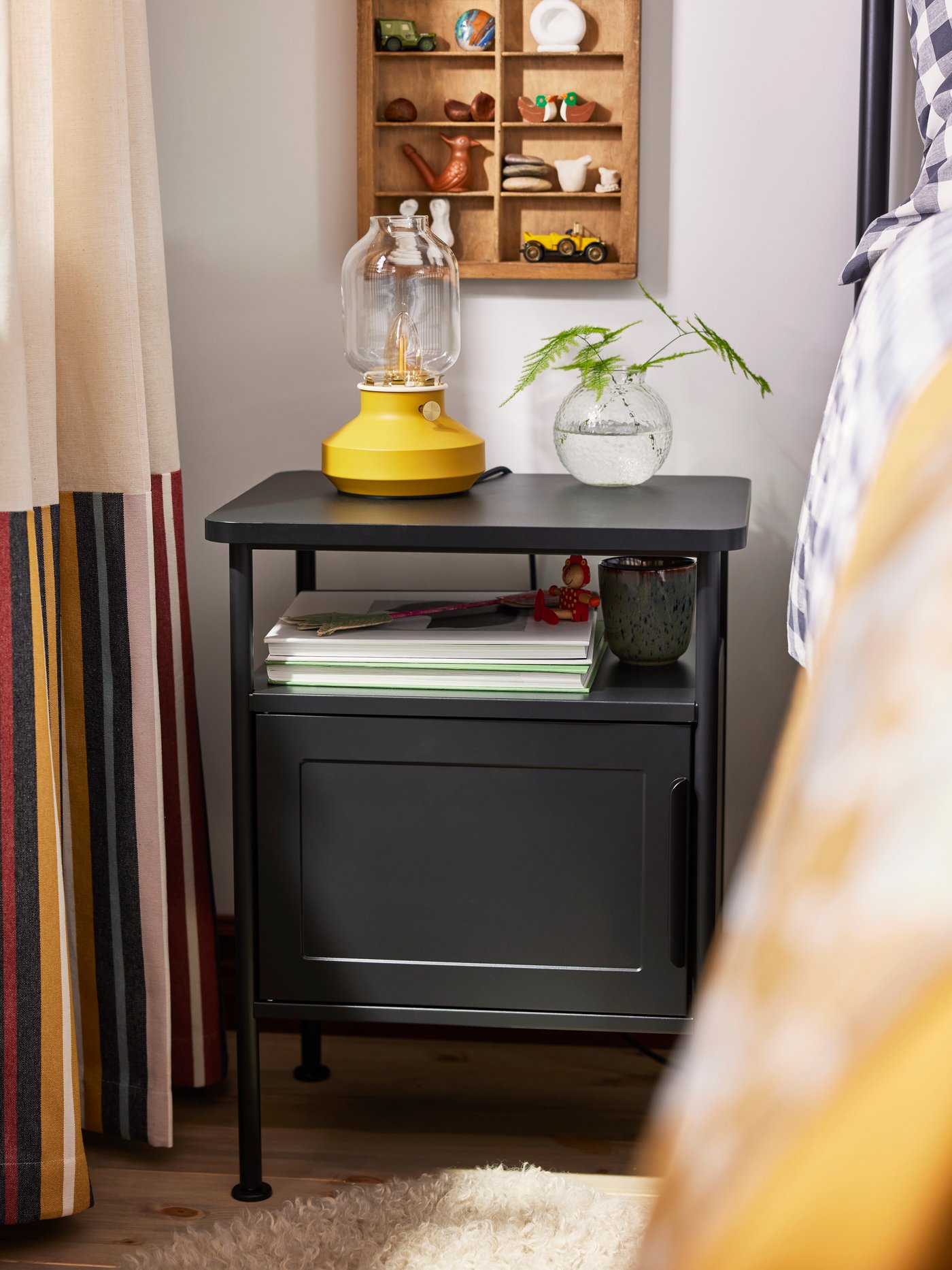 Nahaufnahme des Nachttischs GRÅFJÄLLET in Anthrazit mit gelber Tischleuchte TÄRNABY und kleiner Glasvase darauf.