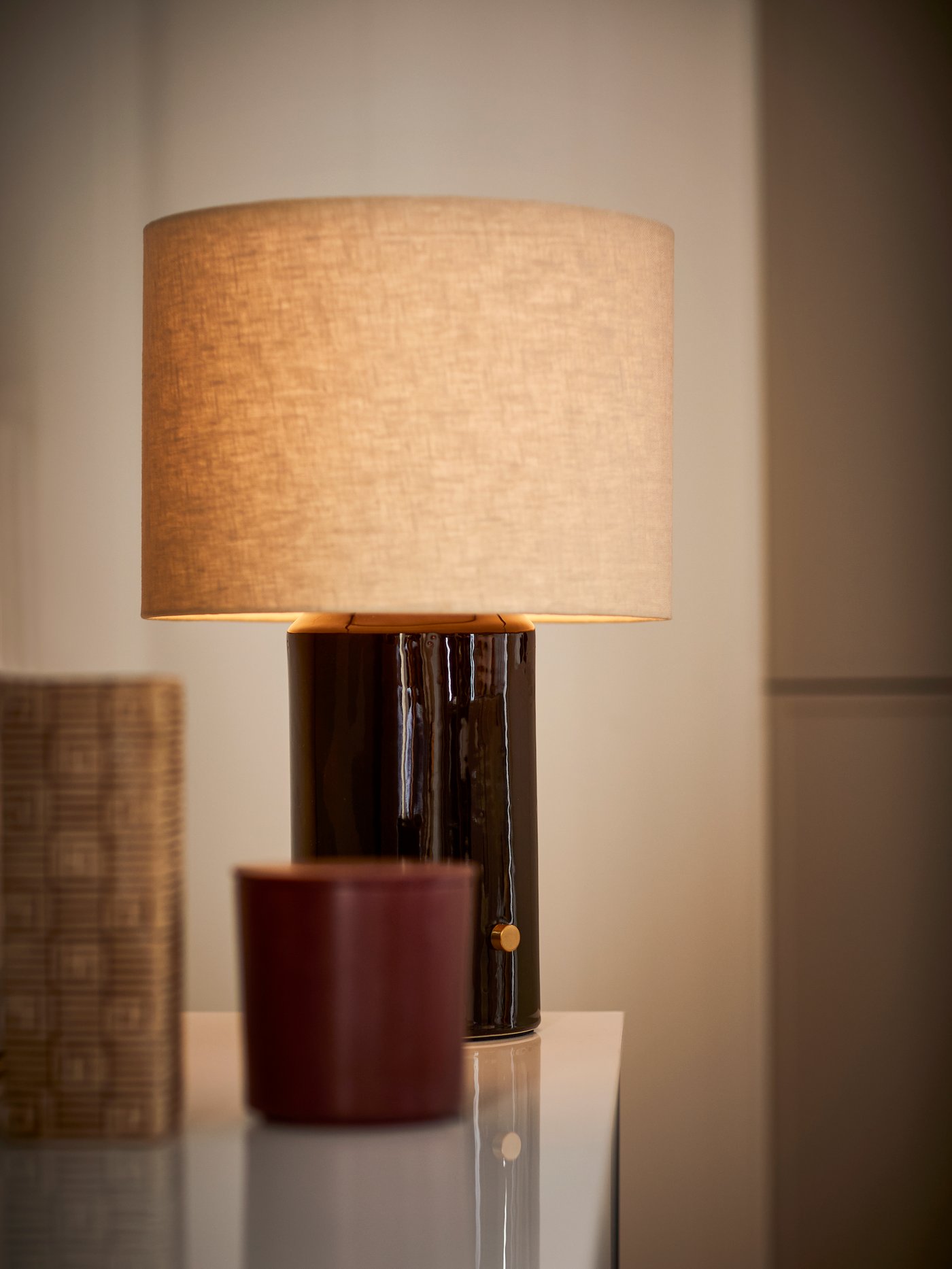 Eine dunkle Tischleuchte steht auf einem weißen Sideboard. Daneben steht ein dunkelrotes Teelichtglas.