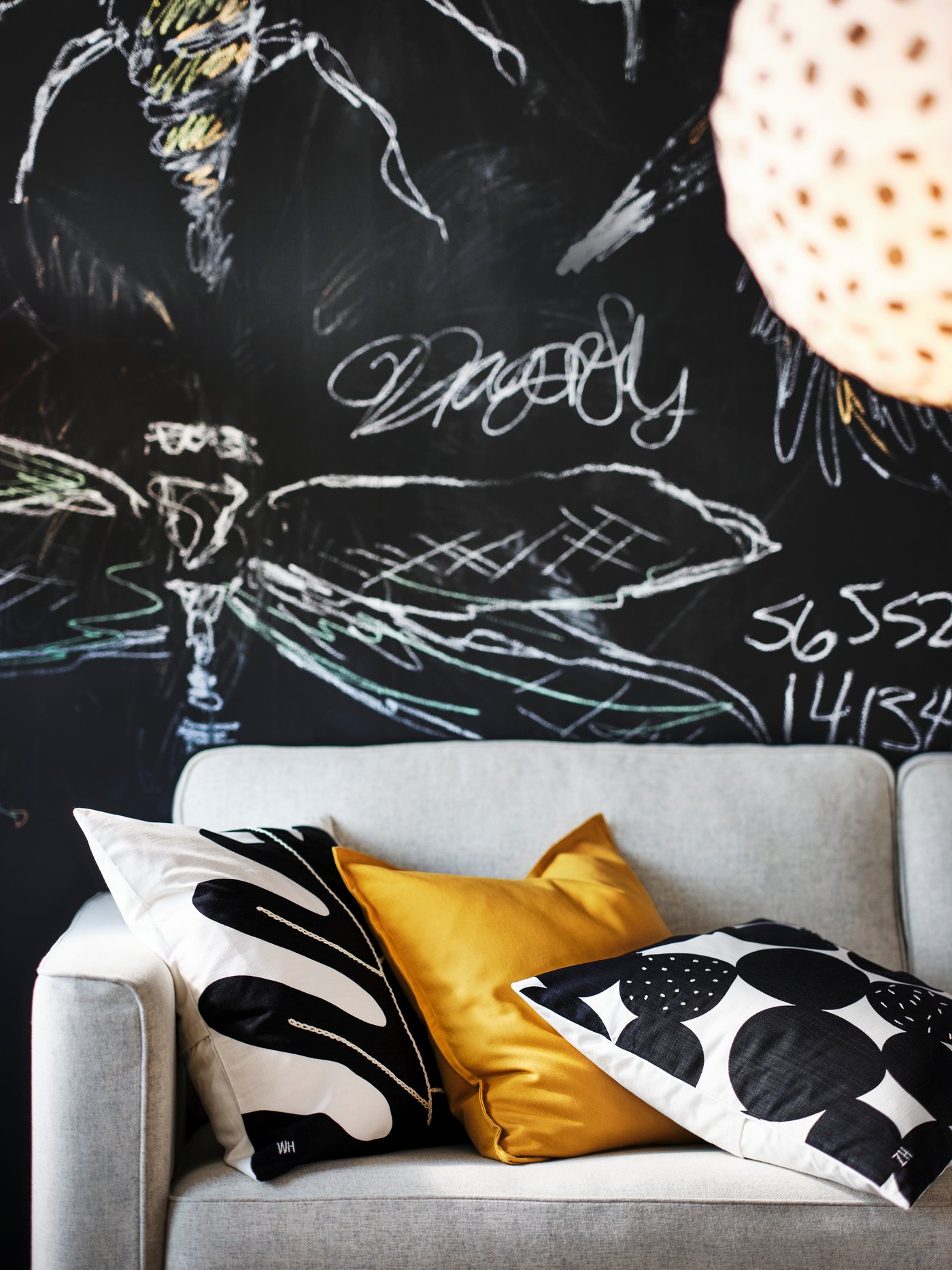 كنبة PÄRUP مغطاة بأغطية وسائد SMÅFROSSÖRT وGURLI. خلفها حائط سبورة سوداء عليها كتابة بالطباشير.