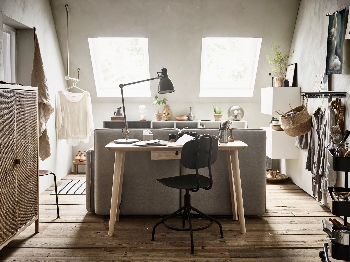 Ljust och mysigt hemmakontor med snedtak och två takfönster som släpper in naturligt ljus. Rummet har trägolv, ett skrivbord med svart stol och en grå soffa bakom. På skrivbordet står en svart lampa, böcker och dekorationer. Till vänster syns ett flätat skåp och hängande kläder, medan hyllor och fler kläder finns till höger om soffan. Inredningen är minimalistisk med naturliga material.