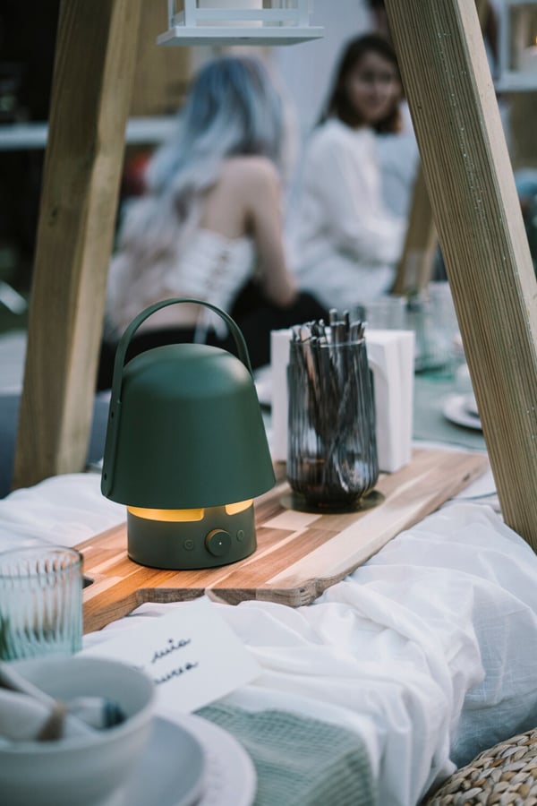 Una tavola bassa allestita e apparecchiata in uno spazio esterno con una lampada/cassa VAPPEBY e un vaso GRADVIS con posate. La tavola è illuminata da lantewrne e luci decorative. Attorno, pouf e cuscini.