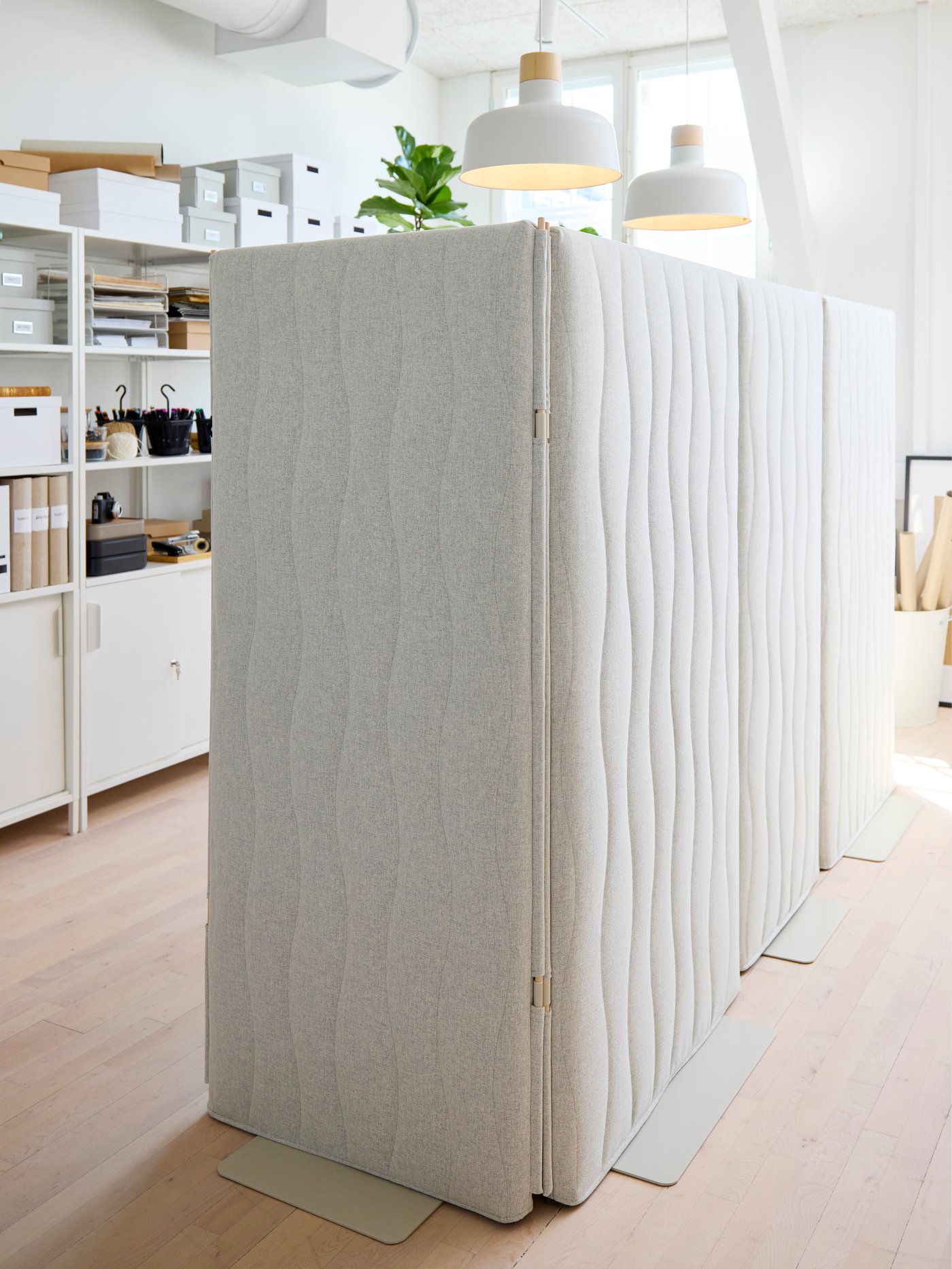 Fristående ljudabsorberande rumsavdelare i ljus textil skapar en avskild arbetsyta i ett modernt kontor. Hyllor med förvaringslådor, växter och takpendlar syns i bakgrunden.