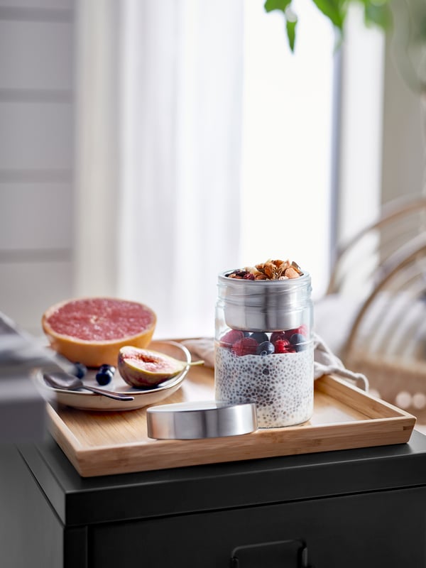 A DAGKLAR jar in clear glass with chia pudding and berries and a steel insert with nuts, beside a plate of fruit on a tray.