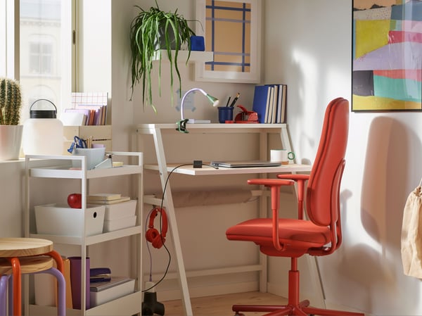 A small corner workspace with a MITTPLAN desk, a red SMÖRKULL office chair, a plant, and a white trolley with storage box