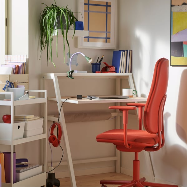 Un piccolo spazio di lavoro ad angolo con una scrivania MITTPLAN, una sedia da ufficio SMÖRKULL rossa, una pianta e un carrello NISSAFORS bianco con contenitore.