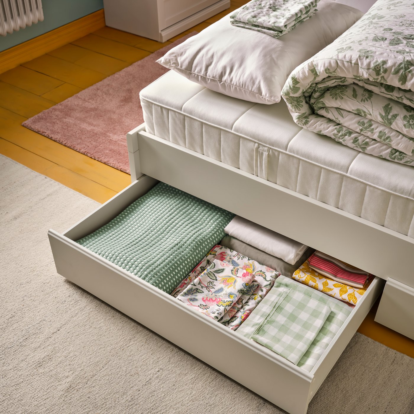 Una recámara con una cama FÅGELFJÄLLET en tono blanco hueso, con cuatro cajas de almacenamiento, arregladas de forma ordenada con la ropa de cama.