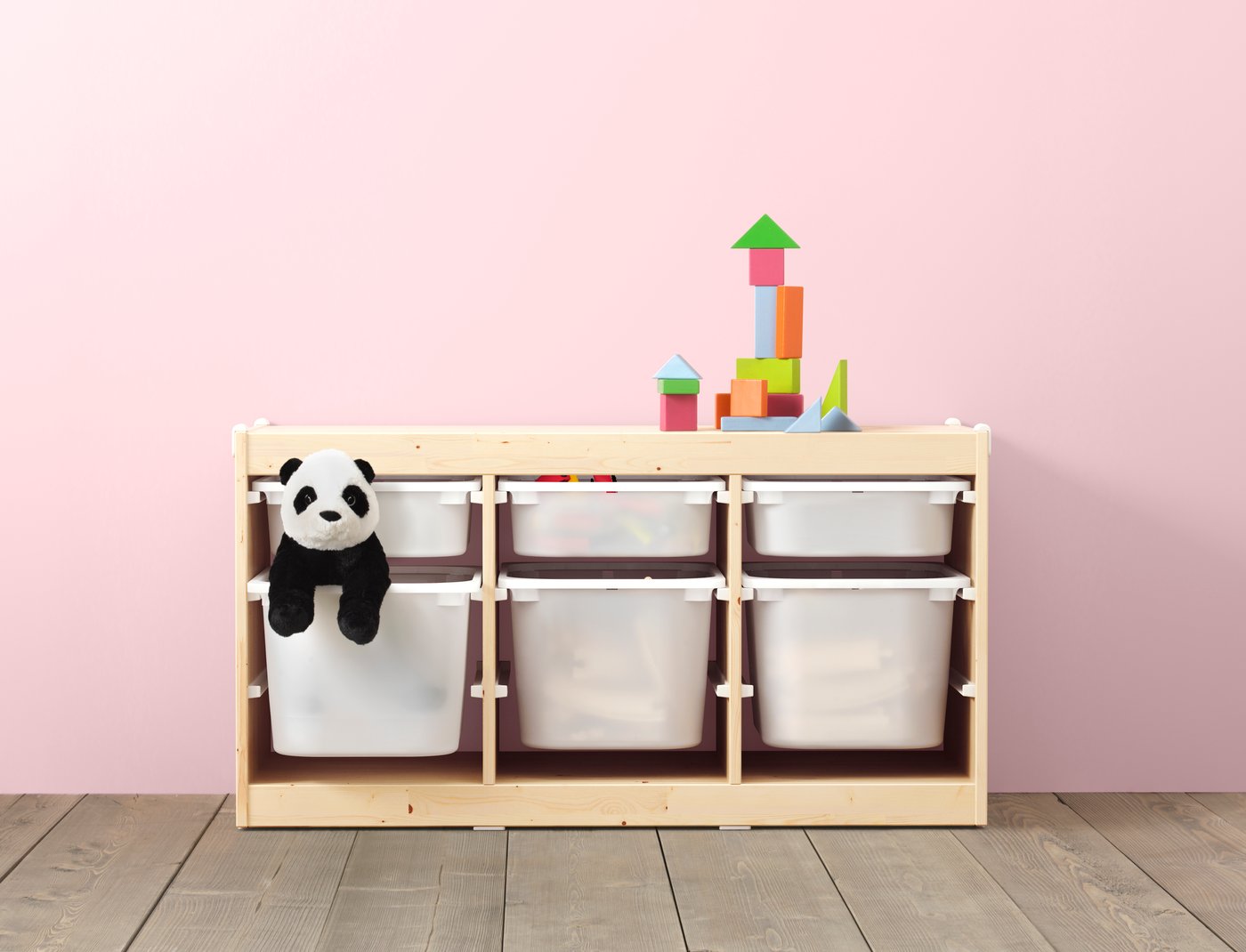 A stained pine TROFAST cabinet with white storage bins. The floor is wooden, and the wall behind it is pink. One of the bins holds a panda plush toy, and a block tower sits on top.