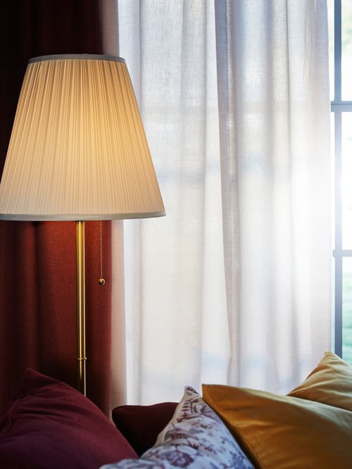 The top half of an illuminated, brass/white ÅRSTID floor lamp is visible between red seating and a curtained window.