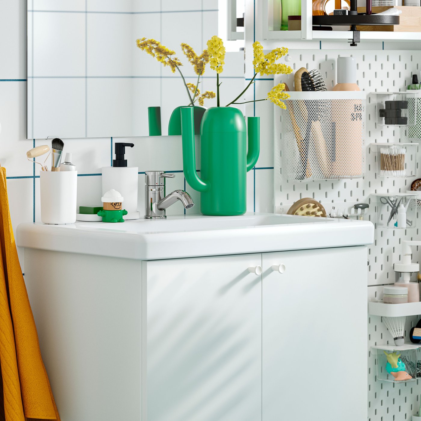 Un armari per a lavabo ENHET blanc amb dues portes, tovalloles de color groc i rosa, un gerro verd i un tauler perforat SKÅDIS amb articles d'higiene personal.