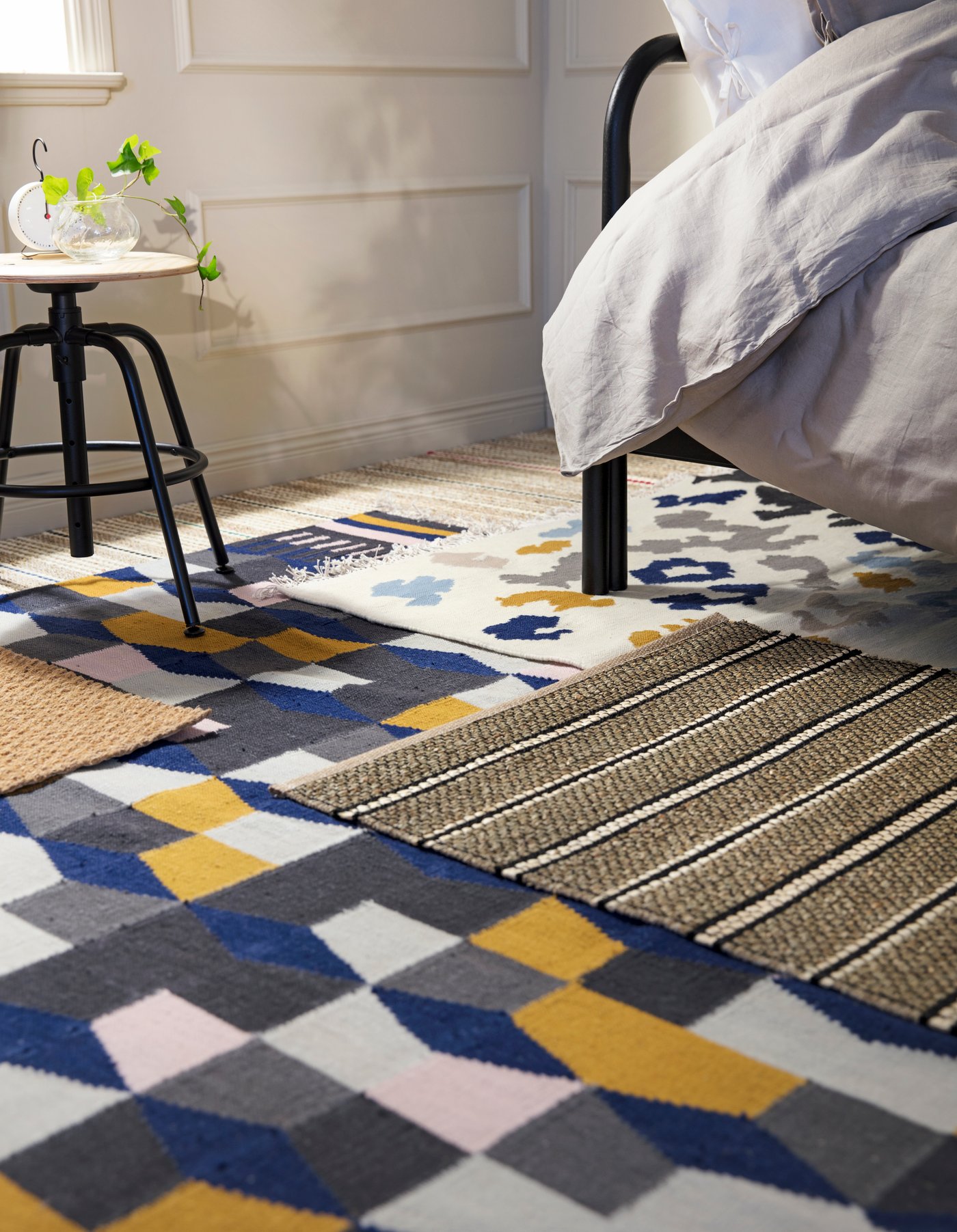 Sovrum med en säng täckt av ett ljusgrått täcke till höger. Golvet är täckt av flera mattor med geometriska mönster i blått, gult, grått och vitt. Till vänster står en KULLABERG stol med svarta ben och gröna växter ovanpå. 