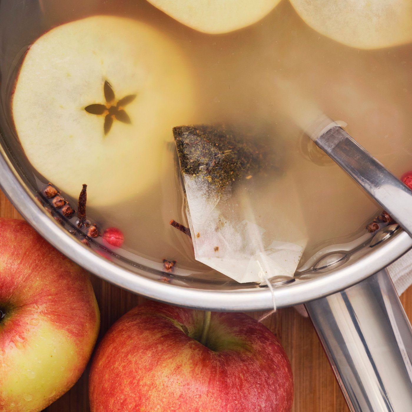 A pot of warm tea with apple slices, spices and a bag of FLÖJT green tea floating and whole apples on the side.