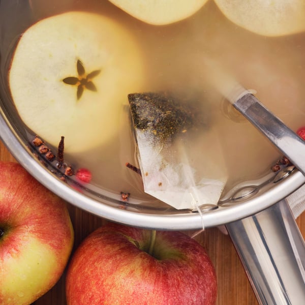 A pot of warm tea with apple slices, spices and a bag of FLÖJT green tea floating and whole apples on the side.
