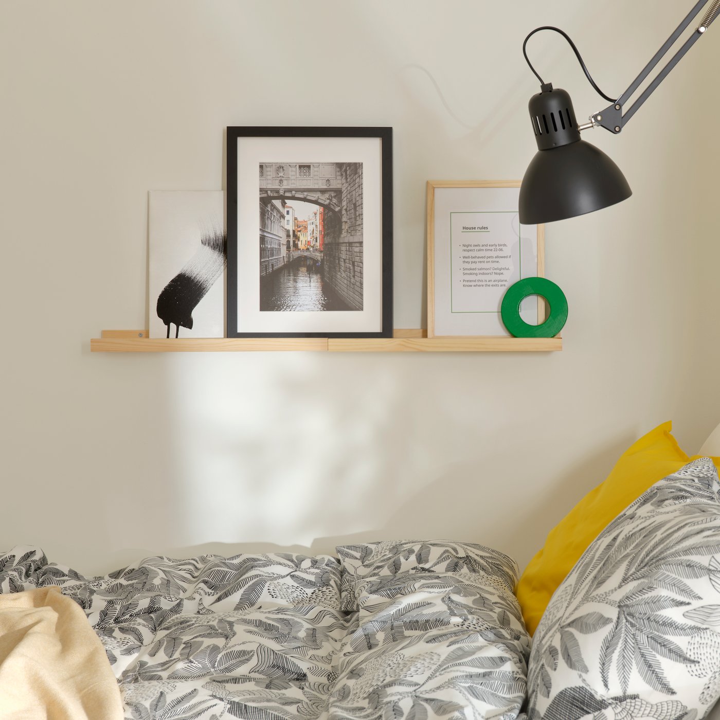 Duas prateleiras para quadros NORDHÄGG em pinho estão numa parede acima da cama, segurando um quadro com as regras da casa e outras obras de arte.