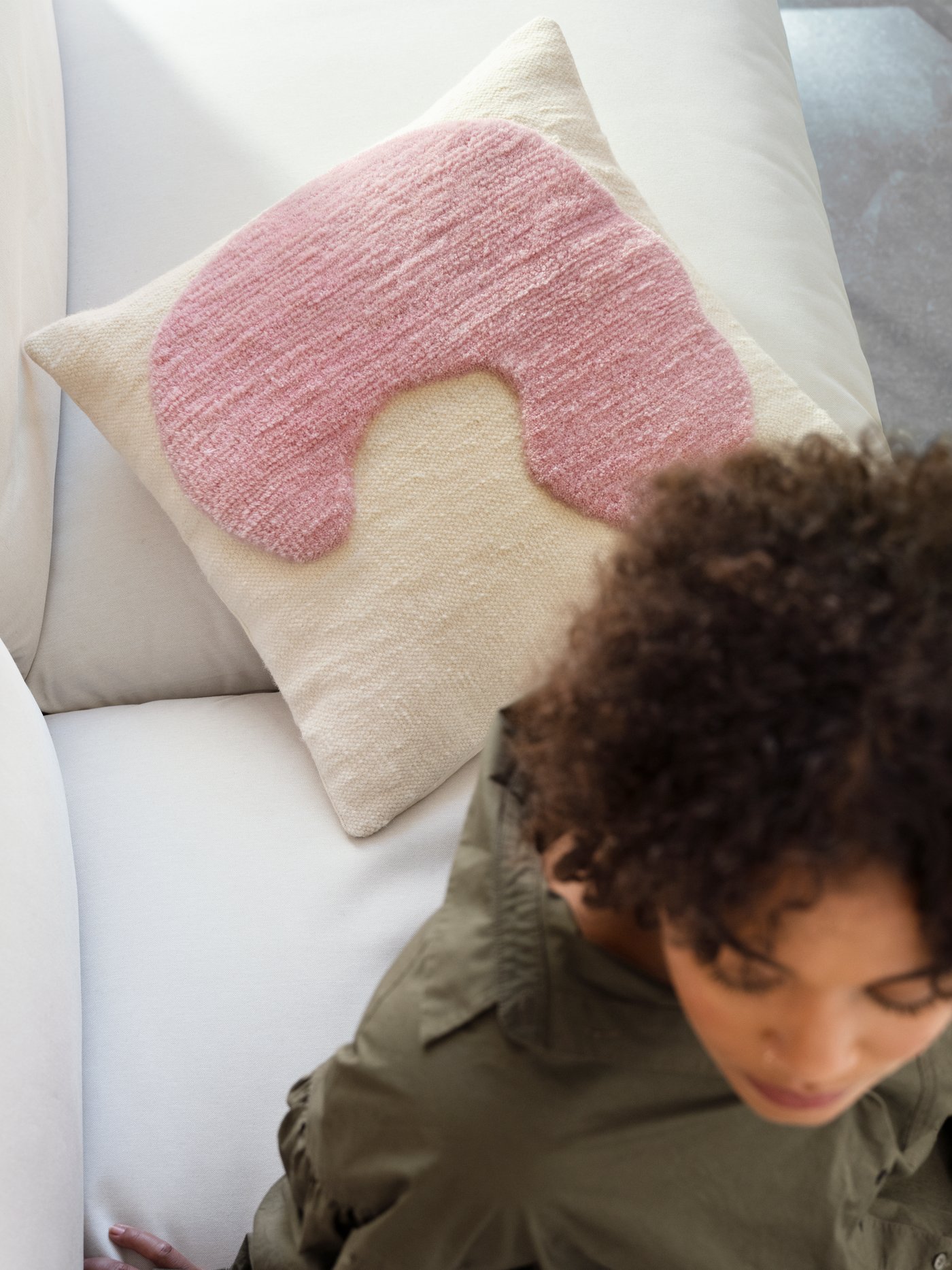 A woman sits in front of a handmade LOKALT cushion that is white with a hand tufted pink patch.