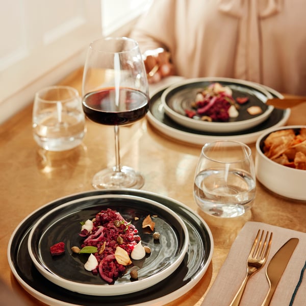 Gröna tallrikar med färgglada sallader serveras på ett ljusbrunt bord med DYRGRIP-glas för vin och vatten.
