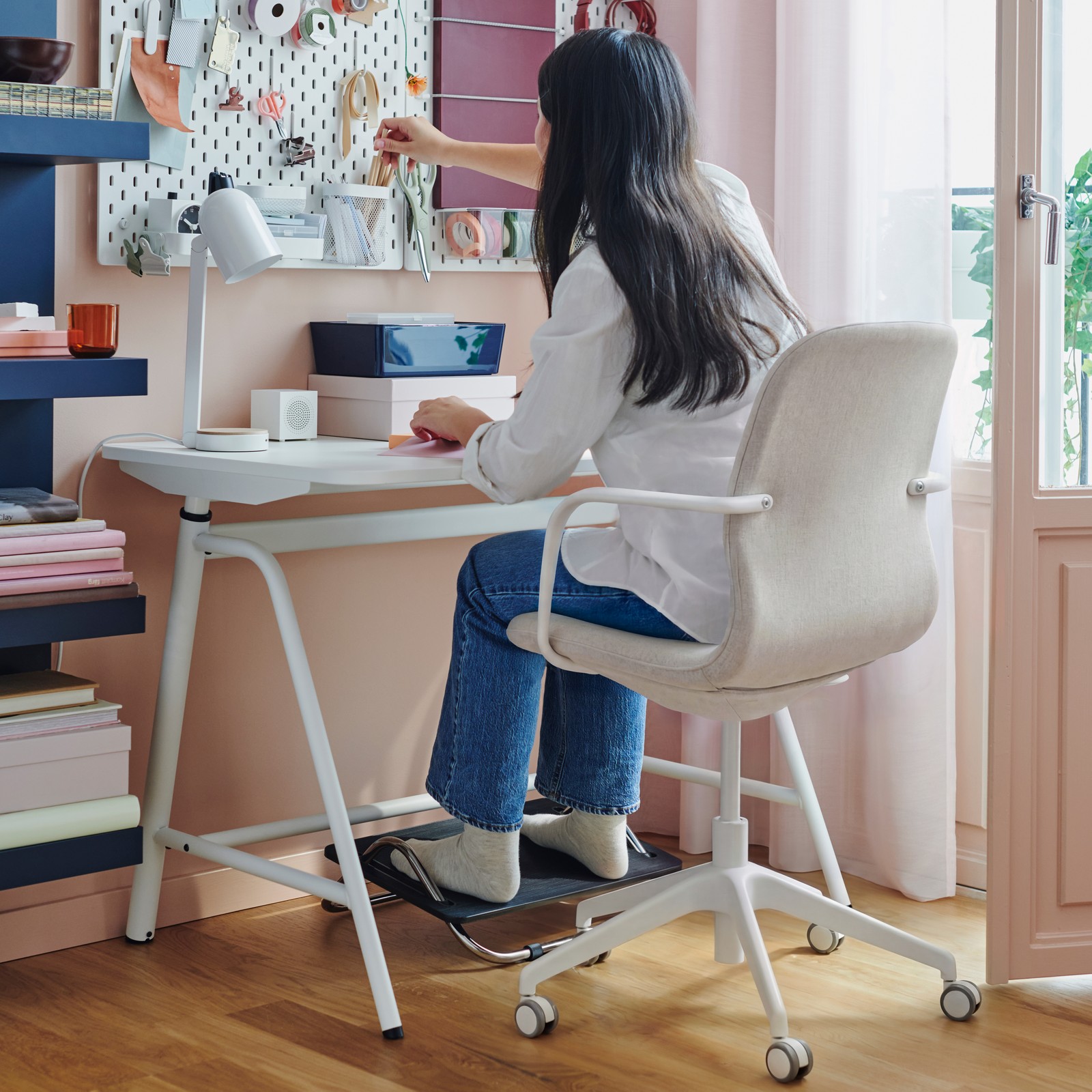 A person sits on a LÅNGFJÄLL conference chair with armrests at a GLADHÖJDEN sit/stand desk with feet on a DAGOTTO foot-rest.