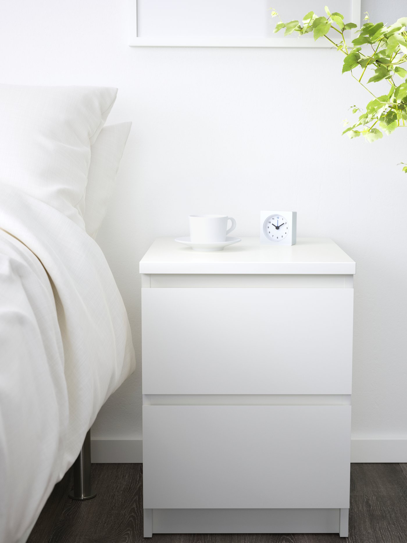 Una cassettiera con 2 cassetti MALM di colore bianco all'interno di una stanza bianca molto luminosa. Sopra, si trova una sveglia e una tazza e accanto un letto.