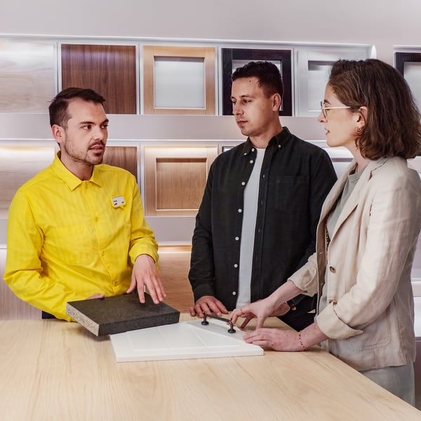 Konsultant IKEA stojący w żółtej koszulce przy stanowisku z frontami kuchennymi i próbkami blatów. Wskazuje na propozycję zestawu i prezentuje go parze klientów.