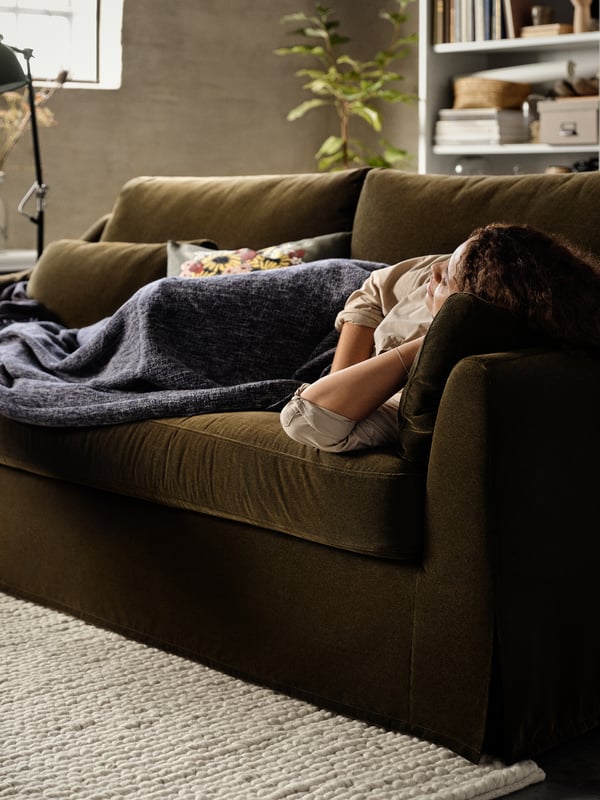 A woman sleeping on a dark olive-green 2-seat sofa, a dark blue throw, cushions, a rug, a white bookcase.