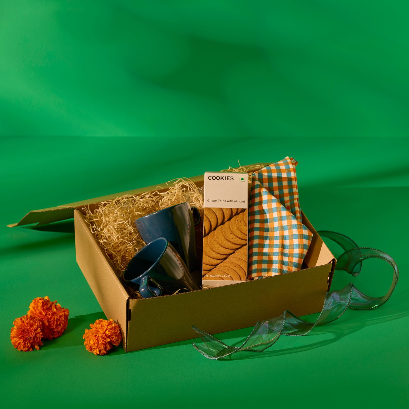 A compact brown gift box with a blue mug, checked napkin, and cookies, decorated with marigold flowers on a green background.