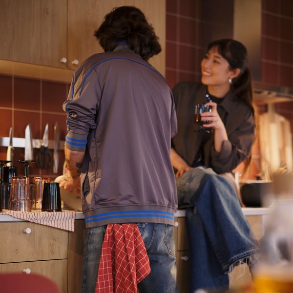 A person sits on a kitchen worktop, looking at another person facing the worktop with a RINNIG tea towel in the back pocket.