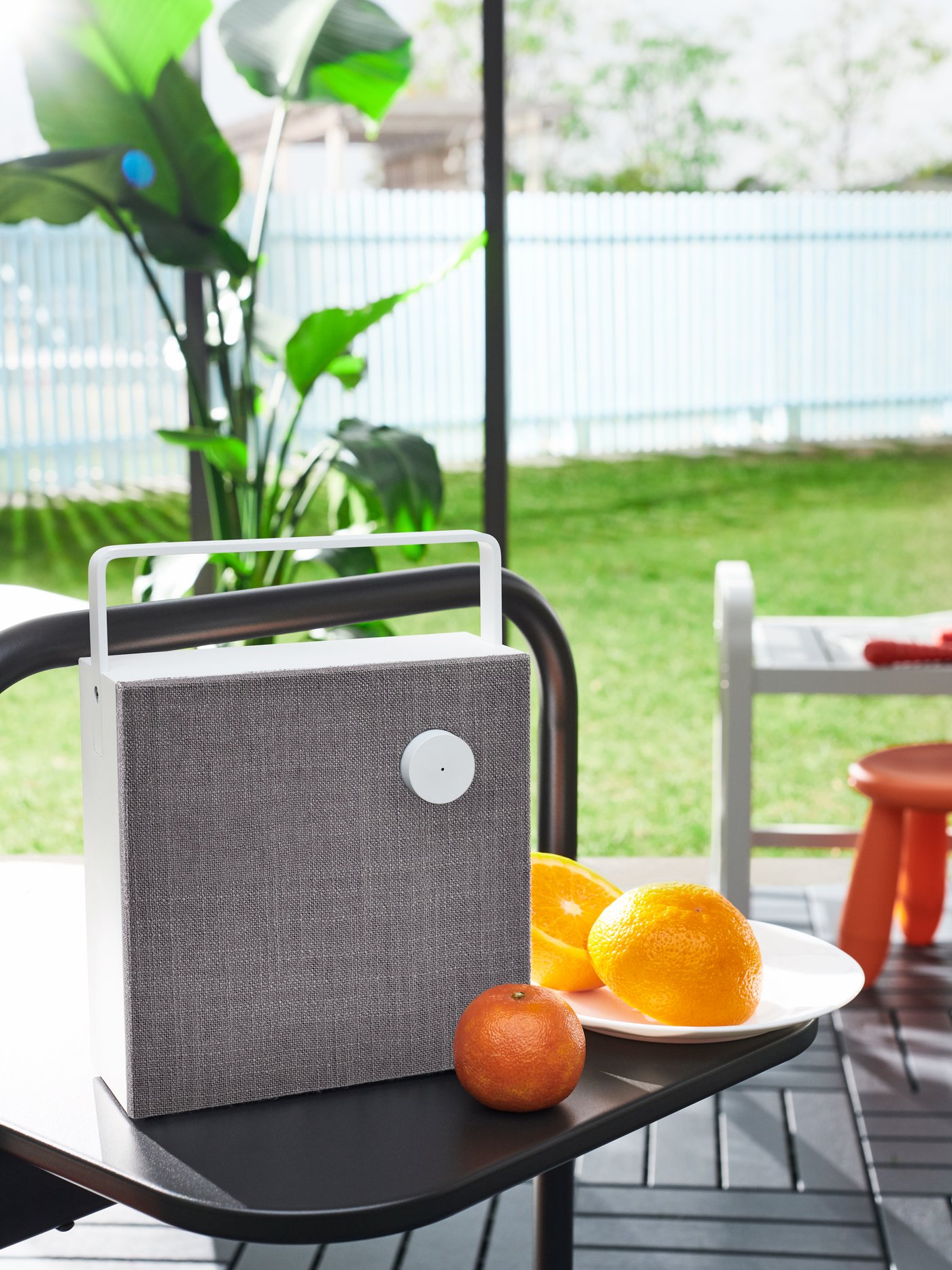 An ENEBY Bluetooth speaker and two oranges sit on a HUSARÖ outdoor side table on some RUNNEN floor decking in a park.