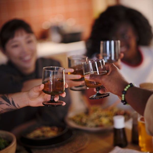 Four people at a table, each holding an IKEA 365+ goblet filled with drinks, reaching towards one another for a toast.