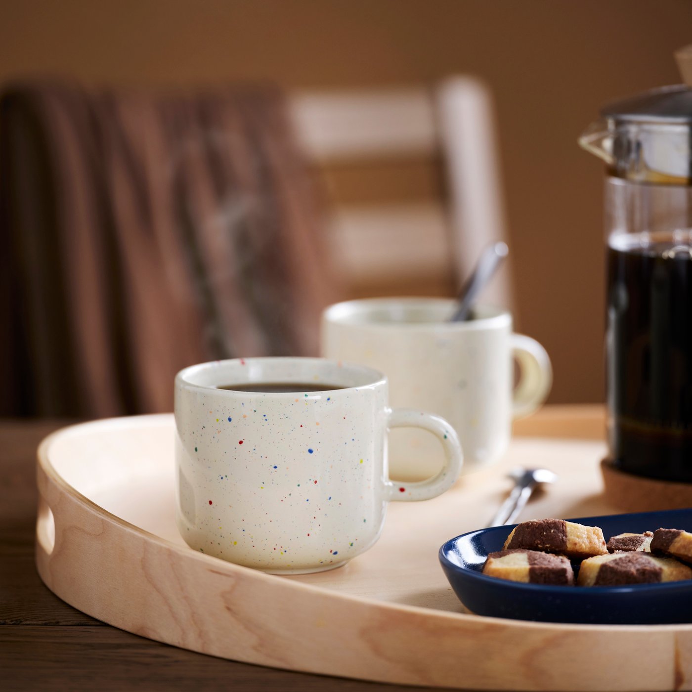 木製トレイの上に、コーヒーが入った斑点模様のセラミックマグカップ2つ、クッキーが盛られた青い皿、スプーン、ガラス製コーヒーポットがテーブルに置かれています。背景には木製の椅子と茶色の布があります。