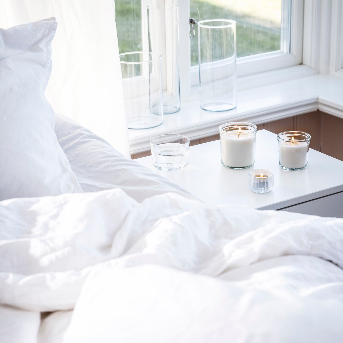 Noční stolek u postele s bílým povlečením, na něm zapálené svíčky IKEA SINNLIG ve skleněných dózách, vedle sklenice vody a prázdné vázy na parapetu.