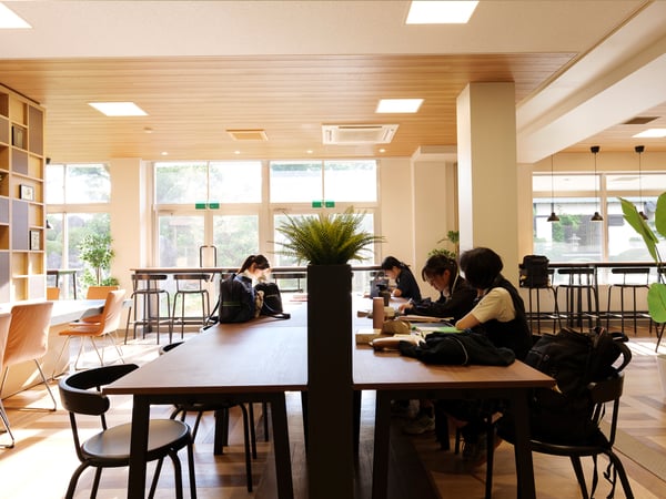 明るい自然光が差し込むカフェ風の共有スペースで、複数の人がテーブルに座り作業や勉強をしている様子