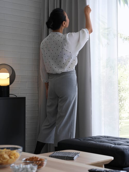 Living room featuring a woman adjusting a pair of curtains in the background, a footstool, side table, and table lamp.