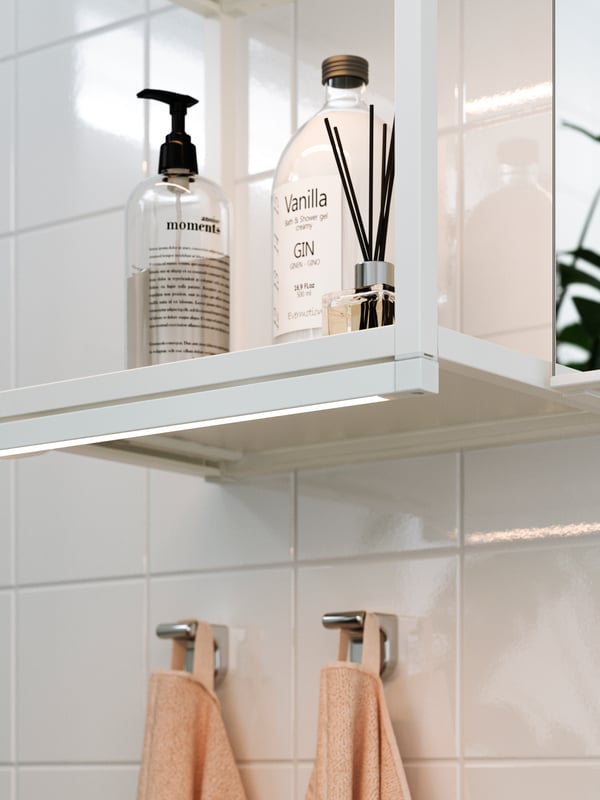 Una barra de iluminación LED SILVERGLANS para baño instalada en la parte inferior de un mueble ENHET blanco y abierto, empotrado a muro con artículos de baño en su interior. Bajo el mueble hay dos ganchos KALKGRUND con toallas de color rosado anaranjado. 