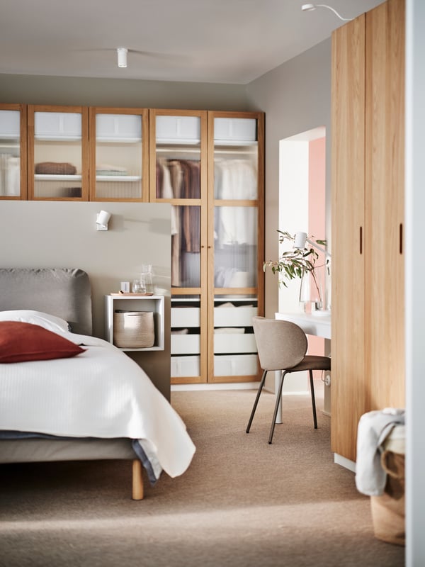 A white SAGESUND upholstered bed covered in white bed linen in front of a PAX/TONSTAD wardrobe in white and oak veneer/glass.