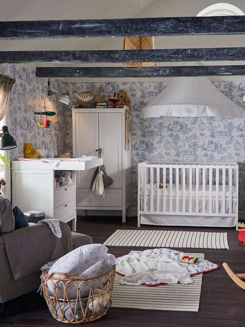 Modern baby room featuring a white cot, a bed canopy, a changing table, a small wardrobe, a basket and an armchair.