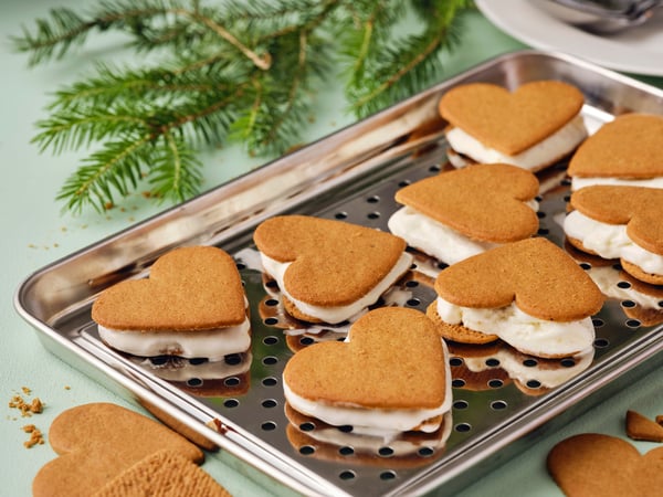 Gelati al pan di zenzero a forma di cuore posti su un vassoio in acciaio uno accanato all'altro. Dietro, un ramo di pino.