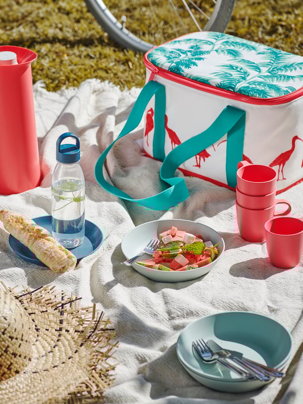 Picnic with a white throw on grass, set with a SOMMARLIV cooling bag, some mugs and a summer salad on a TALRIKA plate.